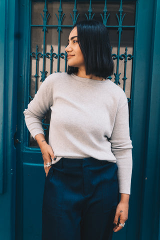 A woman with straight dark hair stands by a teal door, wearing Olive Basics Ellie Reversible Cardigan in TENCEL™ Knit, styled in light gray with dark blue pants, looking to her left.
