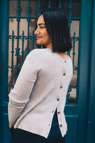 A woman with dark hair stands before a blue door, wearing the Olive Basics Ellie Reversible Cardigan in soft TENCEL™ knit, styled with its signature back buttons.