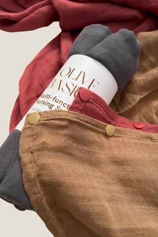 A rolled-up gray Multi-functional Nursing Swaddle by Olive Basics is wrapped in a label and placed on top of overlapping brown and reddish cloths with snap buttons along the edge.