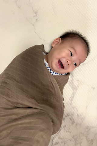 A baby wrapped in the Olive Basics Multi-functional Nursing Swaddle smiles while lying on a light-colored, marble-patterned surface and looking to the right.