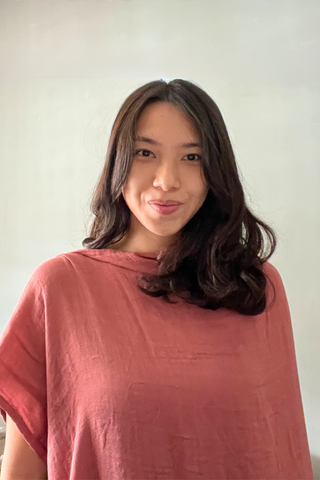 A person with long, wavy dark hair, dressed in a loose, rose-colored top, stands before a neutral background holding the Olive Basics Multi-functional Nursing Swaddle, smiling gently at the camera.