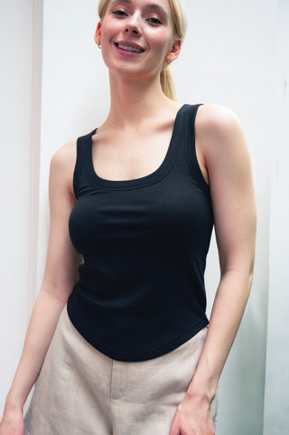 A woman with light hair in a ponytail wears the Olive Basics Anna Reversible Hi-Low Tank in black and beige pants, standing and smiling indoors against a white background.