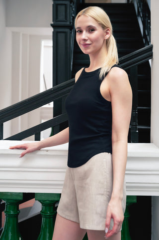 A woman with blonde hair in a ponytail, wearing the Anna Reversible Hi-Low Tank by Olive Basics in black and beige shorts, stands indoors near a staircase and white railing.