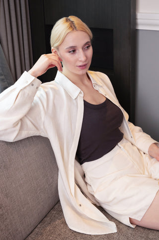 A blonde woman sits on a gray couch, wearing a cream shirt and Olive Basics Joanna Expandable Pocket Shorts in TENCEL™ Linen over a dark tank, looking to the side.