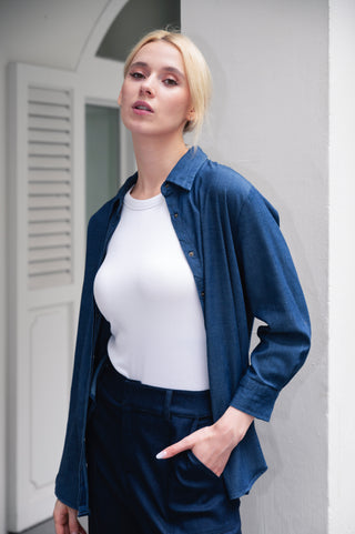 A woman with blonde hair wears the Olive Basics Anna Reversible Hi-Low Tank and a blue button-up shirt with matching pants, standing indoors with one hand in her pocket.