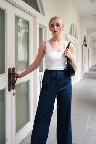 A woman in the Olive Basics Anna Reversible Hi-Low Tank and blue high-waisted pants stands in a hallway, holding a door handle with one hand and carrying a black bag on her shoulder.