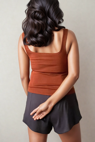 A woman with wavy dark hair wears the Olive Basics Esther Easy Latch Tank in brown with black shorts, standing facing away and crossing her fingers behind her back.