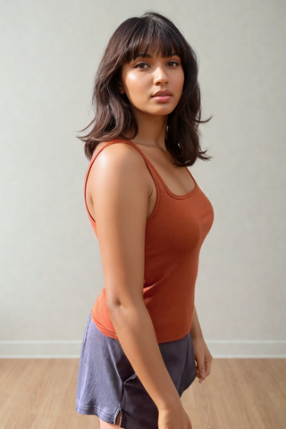 A woman wears the Olive Basics Esther Easy Latch Tank in orange and gray shorts, standing indoors on a wooden floor and looking at the camera with a neutral expression.