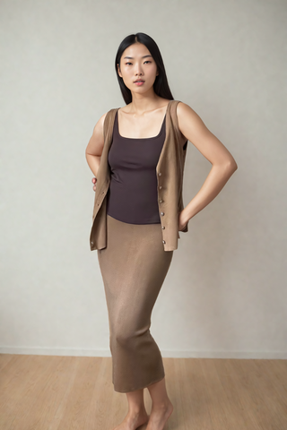 A woman stands indoors against a plain wall, wearing a brown sleeveless vest, a dark brown top, and the Leah Super Stretch Slit Skirt from Olive Basics, with her hands in her vest pockets.