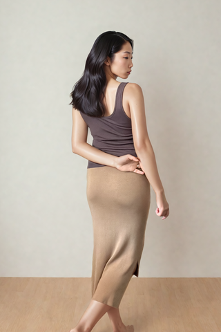 A woman stands barefoot on a wood floor, wearing Olive Basics’ Leah Super Stretch Slit Skirt in tan and a brown tank top, glancing over her shoulder with one arm bent behind her back.