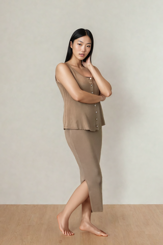 A woman stands barefoot on a wooden floor, wearing the Leah Super Stretch Slit Skirt from Olive Basics—a sleeveless brown buttoned top and matching calf-length breathable skirt—set against a plain light background.