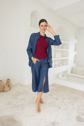 A woman stands barefoot indoors, wearing Olive Basics Delilah All-Occasion TENCEL™ Shirt in blue as a loose layer over a breastfeeding-friendly burgundy top and matching culotte pants. White shelves with ceramics appear in the background.