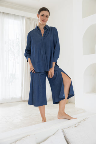 A woman stands indoors wearing the Olive Basics Delilah All-Occasion TENCEL™ Shirt and matching wide-leg culottes with slits, one foot raised on a sofa. The outfit is loose-fitting and breastfeeding friendly.