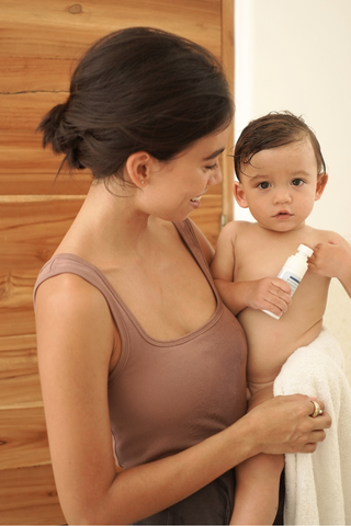 Wearing the Olive Basics Esther Easy Latch Tank, a woman smiles at her baby wrapped in a towel and holding a small bottle; they share a joyful moment together in a cozy bathroom.