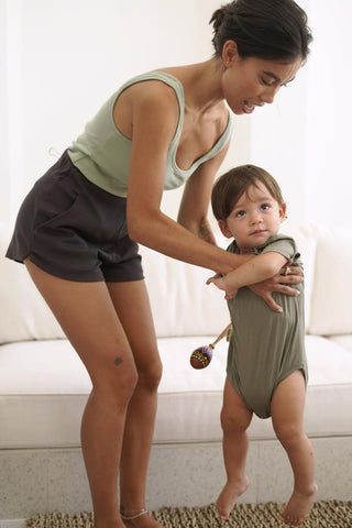 An adult supports a toddler standing on a couch. The toddler, wearing Olive Basics Rebecca Expandable Pocket Shorts and holding a small rattle, looks ahead as the adult steadies them under the arms.
