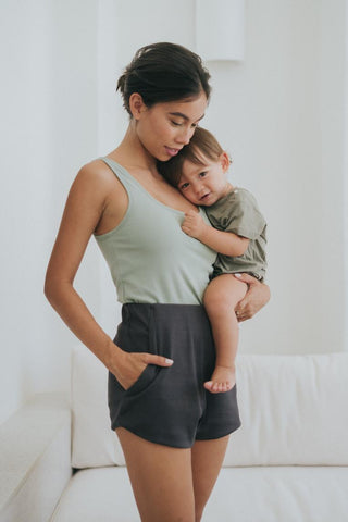 A woman stands indoors holding a toddler on her hip. She wears Olive Basics Rebecca Expandable Pocket Shorts with an adjustable waistband; both are in casual attire and have neutral expressions.