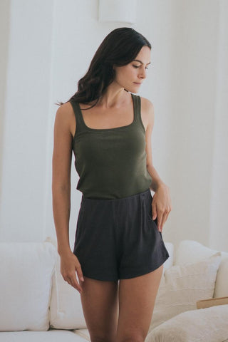 A woman stands indoors wearing Olive Basics Rebecca Expandable Pocket Shorts—black with an adjustable waistband—paired with a sleeveless olive green top, one hand on her leg and the other on her hip.