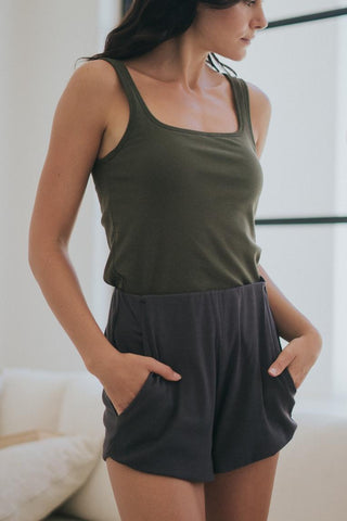 A woman stands indoors with her hands in the pockets of Olive Basics’ Rebecca Expandable Pocket Shorts, paired with a sleeveless olive green top and featuring an adjustable waistband.