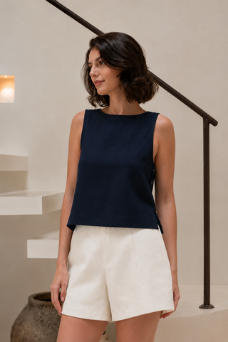 A woman stands indoors in front of a staircase with a neutral-colored background, wearing the Olive Basics Leigh-Anne Reversible Button Top in Midnight Blue and white shorts, which offers convenient breastfeeding access.