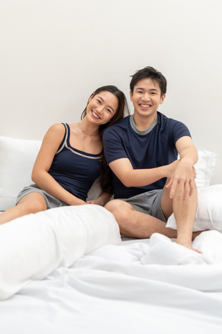Two people in casual sleepwear, including the Noah Lightweight Multi-functional Tee by Olive Basics—crafted from a TENCEL™ wool blend—sit on a white bed, smiling at the camera against a plain background. Their moisture-wicking attire promises comfort and ease for a restful night.