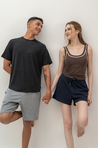 Two people stand against a white wall, holding hands and smiling at each other. Both are dressed in casual shorts and tops, including the Olive Basics Noah Lightweight Multi-functional Tee—crafted from a TENCEL™ wool blend for exceptional moisture-wicking comfort.