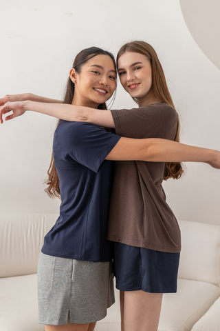 Two women stand facing each other with arms around each other, smiling at the camera in a casual indoor setting. Both are dressed comfortably—one wears the Chloe Relaxed Croppable Tee by Olive Basics, while the other sports a soft lounge set crafted from TENCEL™ Wool Spandex—blending effortless style and cozy relaxation.