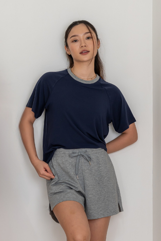 A woman stands against a white wall, wearing Olive Basics’ Chloe Relaxed Croppable Tee in navy—a comfortable top from the lounge set—paired with soft light gray drawstring shorts made from TENCEL™ Wool Spandex, her left hand resting on her hip.