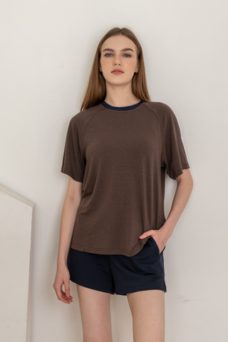 A woman with long hair wears the Olive Basics Chloe Relaxed Croppable Tee—a brown short-sleeve lounge top made from soft TENCEL™ wool spandex. She pairs it with navy shorts and stands against an off-white wall, one hand casually in her pocket.