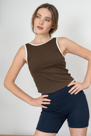 A woman poses with hands on hips, wearing the Olive Basics Baby Anna Reversible Crop Tank in Dirty Mocha and navy shorts, standing against a plain light-colored background.