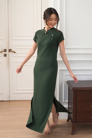 A woman stands barefoot in a room with white walls and wooden furniture, wearing the Eliza Super Stretch Cheongsam Dress by Olive Basics—a dark green, short-sleeved, floor-length dress with side slits.