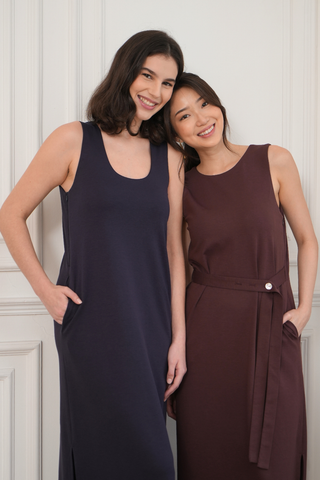 Two women smile indoors, each with hands in pockets. One wears a navy dress, the other a maroon Noel Reversible Multi-way Maxi Dress by Olive Basics, featuring a stylish reversible neckline.