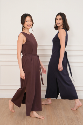 Two women stand barefoot indoors against a white wall, wearing Olive Basics' Noel Reversible Multi-way Maxi Dress—one in maroon, one in navy.