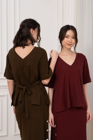 Two women stand indoors; one faces away in a brown ribbed top and skirt, while the other smiles at the camera wearing Olive Basics’ Tabitha Reversible Multi-way Tee (TENCEL Cotton Rib) in burgundy with a V-neck and reversible neckline.