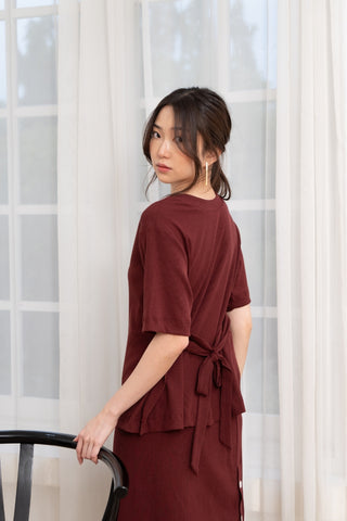 A woman in the Olive Basics Tabitha Reversible Multi-way Tee (TENCEL Cotton Rib) stands by a black chair, looking over her shoulder in front of sheer white curtains and a window.