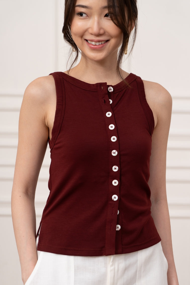 A woman wearing the Olive Basics Grace Reversible Button Tank in maroon with white buttons and white pants stands indoors, smiling.