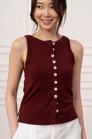 A woman wearing the Olive Basics Grace Reversible Button Tank in maroon with white buttons and white pants stands indoors, smiling.