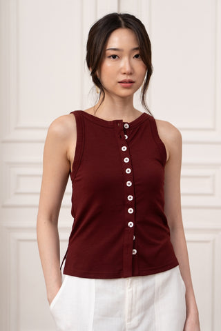 A woman stands in front of a light-paneled wall wearing the Olive Basics Grace Reversible Button Tank in burgundy with white buttons, paired with white pants.