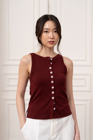 A woman wears the Olive Basics Grace Reversible Button Tank in maroon with white buttons and white pants, standing against a light-colored paneled background.