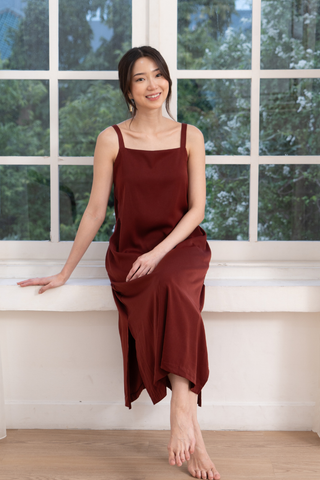 A woman in the Olive Basics Hannah Reversible Midi Dress in wine sits barefoot on a white windowsill, smiling at the camera with trees visible through the window behind her.
