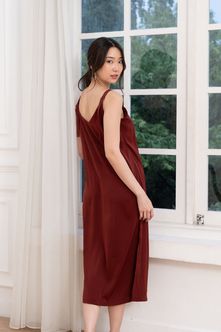 A woman stands indoors by a large window, looking over her shoulder at the camera in the Olive Basics Hannah Reversible Midi Dress.