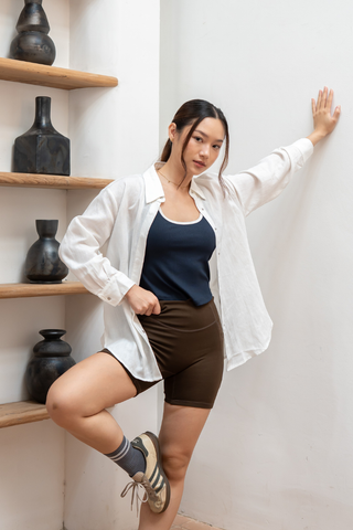 A woman wearing a white shirt, brown shorts, and the Olive Basics Baby Anna Reversible Crop Tank in navy Modal Cotton Rib with a built-in shelf-bra poses indoors against a white wall beside shelves displaying dark ceramic vases.