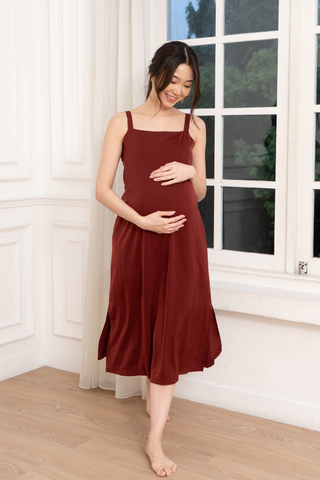 A pregnant woman smiles as she stands barefoot by a large window, gently holding her belly while wearing the Olive Basics Hannah Reversible Midi Dress.