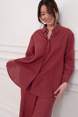 A woman in the Marie Everyday Shirt in TENCEL™ Linen by Olive Basics, paired with matching pants, stands against a white wall smiling and holding out her shirt to display its structured drape.