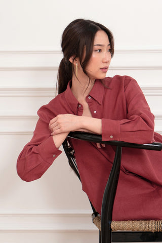 A woman with long dark hair in a ponytail wears the Olive Basics Marie Everyday Shirt in TENCEL™ Linen, Rose Taupe, sitting sideways on a black chair and looking left against a white wall background.