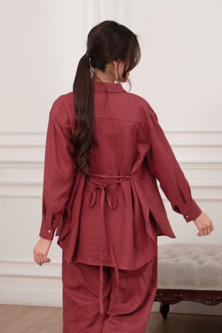 A person with long dark hair in a ponytail wears the Olive Basics Marie Everyday Shirt in TENCEL™ Linen, styled loosely and tied at the waist. They are seen from behind, standing in a white room with panelled walls.