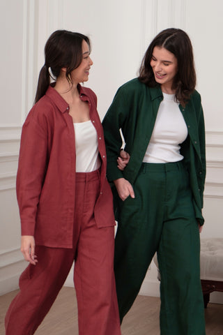 Two women indoors, smiling—one in a red outfit styled with the Olive Basics Marie Everyday Shirt in TENCEL™ Linen, the other in green with a white top—both radiate effortless style and warmth.