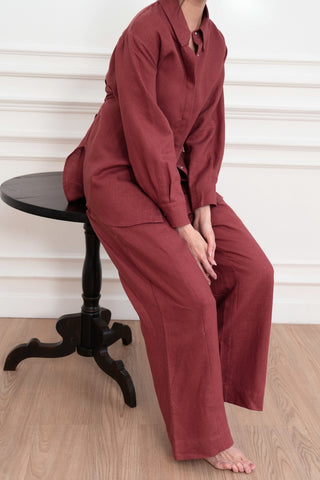 A person in Olive Basics’ Jesse Wide-leg Pocket Pants in TENCEL™ Linen, styled loosely in maroon, sits barefoot on a black wooden chair against a paneled white wall.