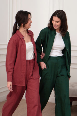Two women share a smile while walking indoors. One wears a red shirt and the Olive Basics Jesse Wide-leg Pocket Pants in TENCEL™ Linen; the other wears a green shirt and pants. Both have white tops underneath.