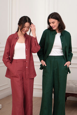 Two women stand indoors in matching Olive Basics Jesse Wide-leg Pocket Pants in TENCEL™ Linen—one pair in red and one in dark green—styled with button-up shirts, white tops underneath, and adjustable waistbands for comfort.
