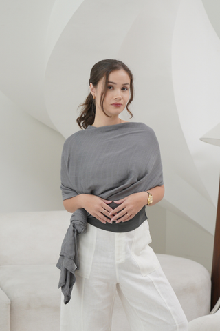 A woman in a gray shawl and white pants stands indoors, hands clasped, against white curved decor, evoking the gentle comfort of Olive Basics' Multi-functional Nursing Swaddle.